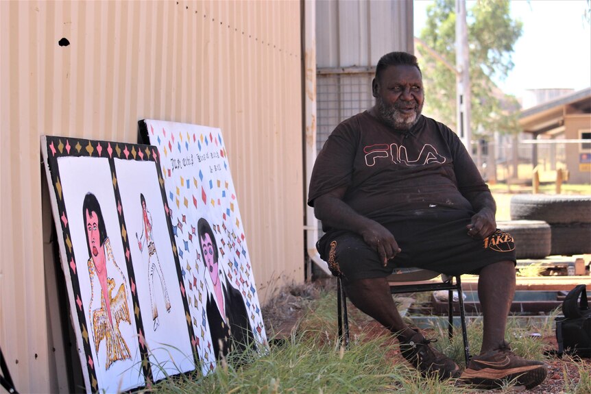 He sits on a chair outside with two Elvis paintings to his right