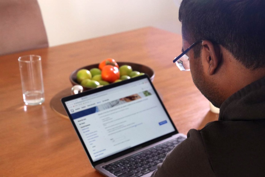 A man pictured from behind, sitting at a kitchen table looking at an open laptop screen.