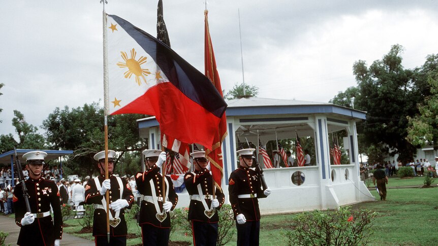 On an overcast day, you view American soldiers holding rifles, the Philippine, and American flags in an official ceremony.