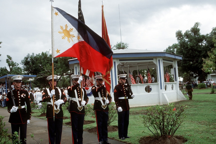 On an overcast day, you view American soldiers holding rifles, the Philippine, and American flags in an official ceremony.