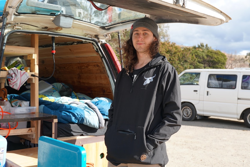 A young man in a beanie and black jacket stands in the sun by the back of a camper van.