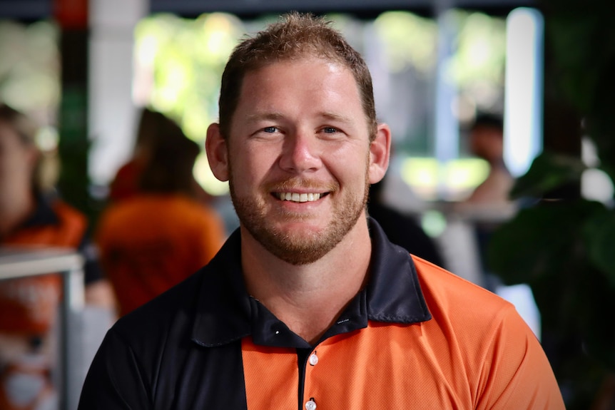 A man in a orange collared shirt smiles into the camera. 