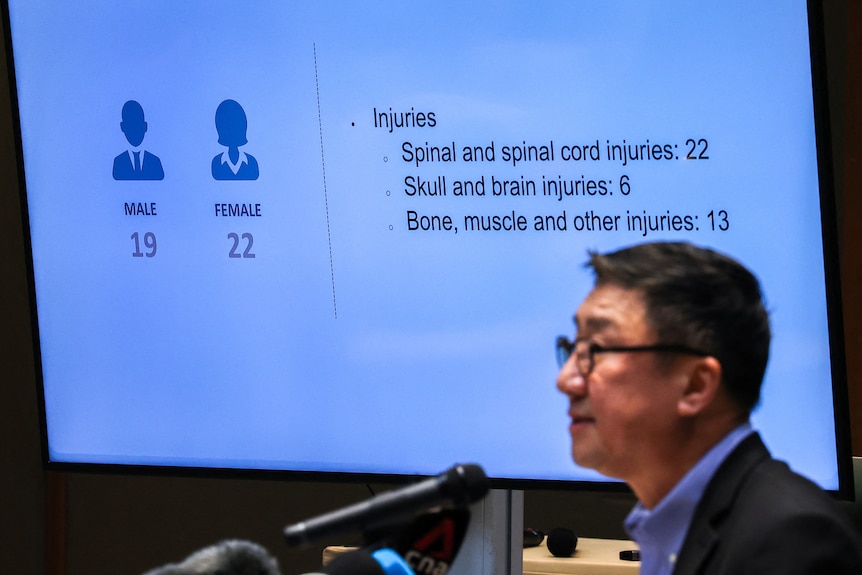 A middle-aged man with dark hair and glasses speaks in front of a screen displaying statistics about airline passenger injuries.