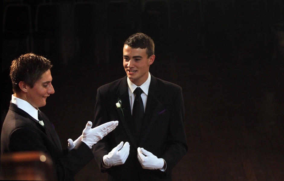 Two young men in tuxedos and white gloves talk.