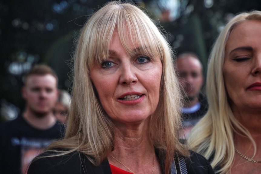A close-up shot of an older woman with blonde hair looking to her right, with another blonde woman on her left.