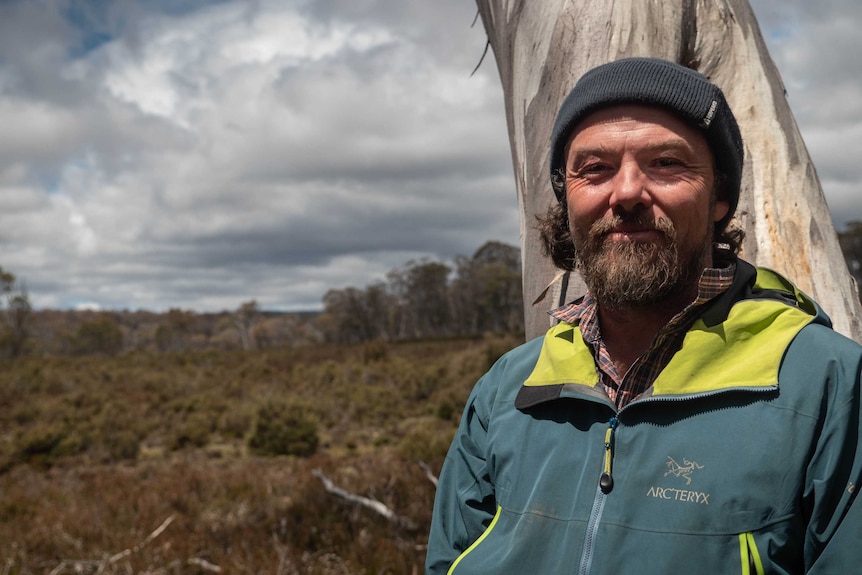Dan Broun has a rugged beard, raincoat and beanie. He's standing in front of a white gum tree trunk with an open plain behind.