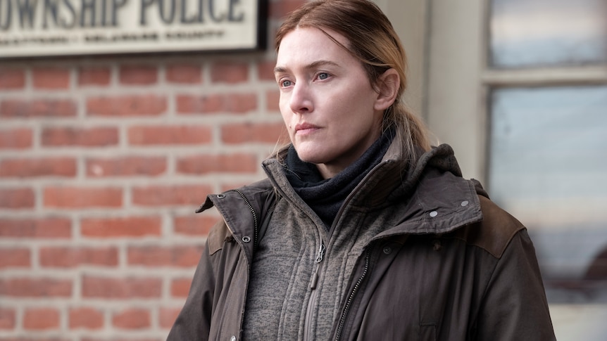 Kate Winslet as an exhausted detective standing outside a police station in tv series Mare of Easttown