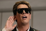 Milo Yiannopolous raises his hand to his hear during an event in Parliament House. He's wearing sunglasses despite being inside.