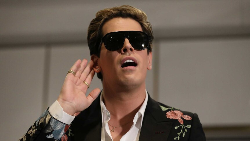 Milo Yiannopolous raises his hand to his hear during an event in Parliament House. He's wearing sunglasses despite being inside.