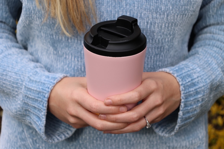 A woman's hands holding a pink and black coffee cup, wearing a blue jumper