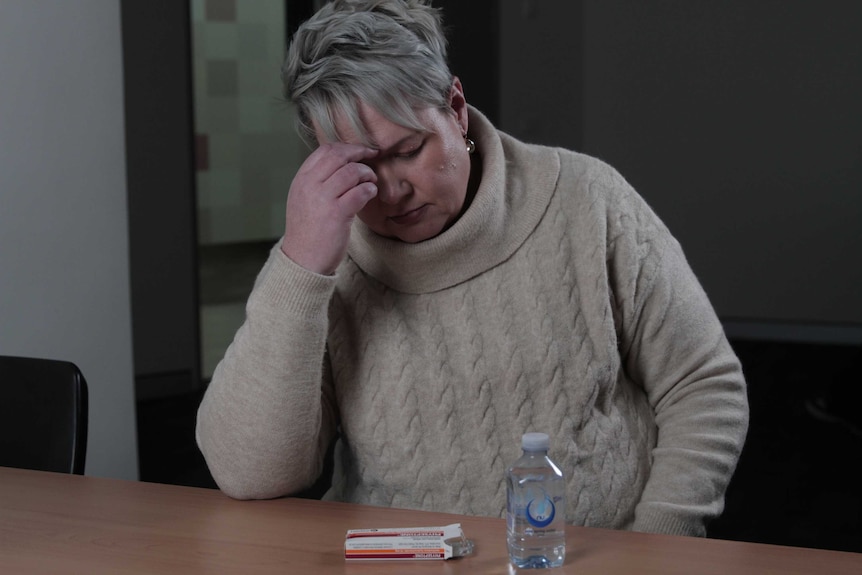 Jodie Carter sitting with her head in her hand looking at her last packet of pain killers which are on the table some water