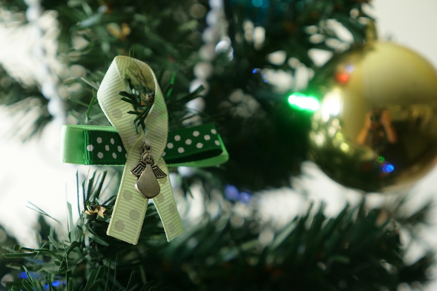 A green ribbon with a small silver angel charm in the middle hanging on a Christmas tree.