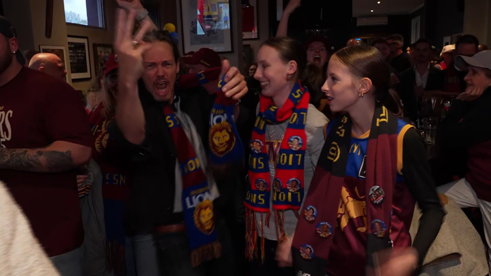 People in a dark bar, many in Brisbane Lions supporter gear, with one man holding three fingers in the air.