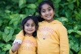 Two young girls, both wearing jumpers that say "kindness counts" smile with their arms around each other.