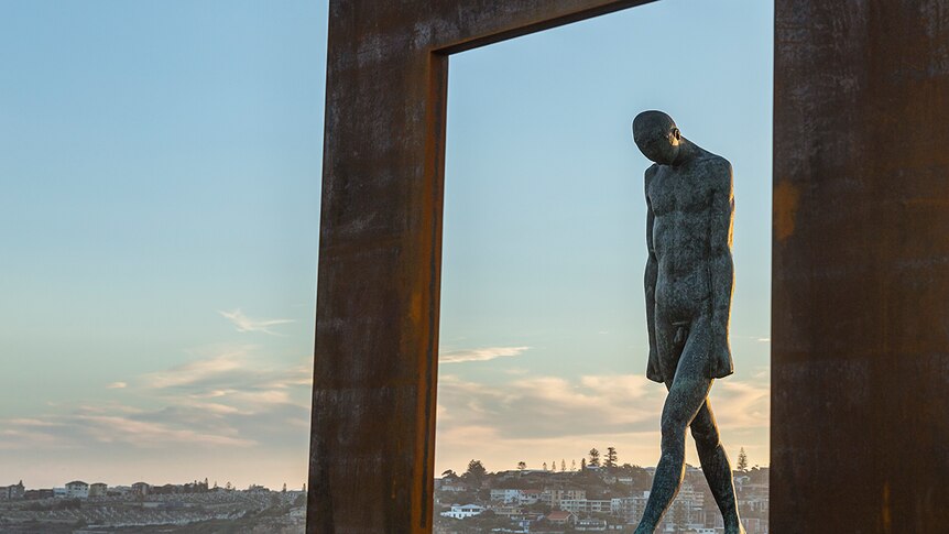 Image of two metal sculptures: a walking man, head down, framed by a 'window frame-like structure'. Blue afternoon sky behind.