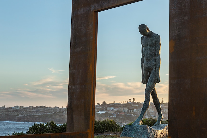 Image of two metal sculptures: a walking man, head down, framed by a 'window frame-like structure'. Blue afternoon sky behind.