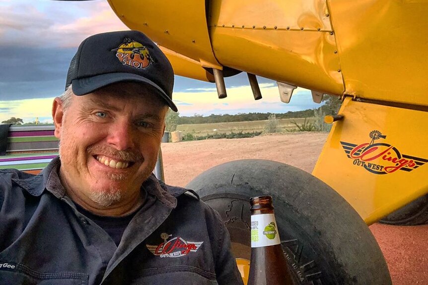 A man with a grey beard smiles in a cap and worn grey shirt with a beer in front of a yellow plane outside.