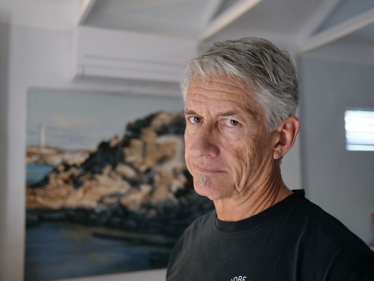 A close up of Keiran wearing a black t-shirt, looking at the camera inside a house, with a painting of a lighthouse behind.