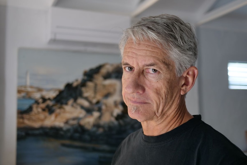 A close up of Keiran wearing a black t-shirt, looking at the camera inside a house, with a painting of a lighthouse behind.