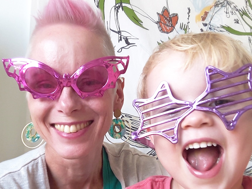 A woman with pink hair and pink sunglasses smiles. A young boy is wearing purple sunglasses with a cheeky smile on his face
