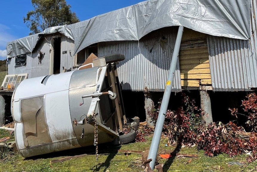 A trailer lies on its side in front of a corrogated iron building with a tarp on its roof.