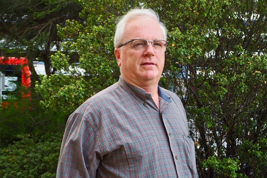 A man with glasses and a check shirt stands in front of a tree.