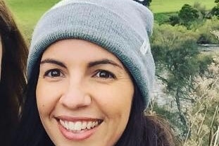 A woman smiles for a selfie, wearing a beanie, scarf and coat in a hillside location, taking a break from social work.