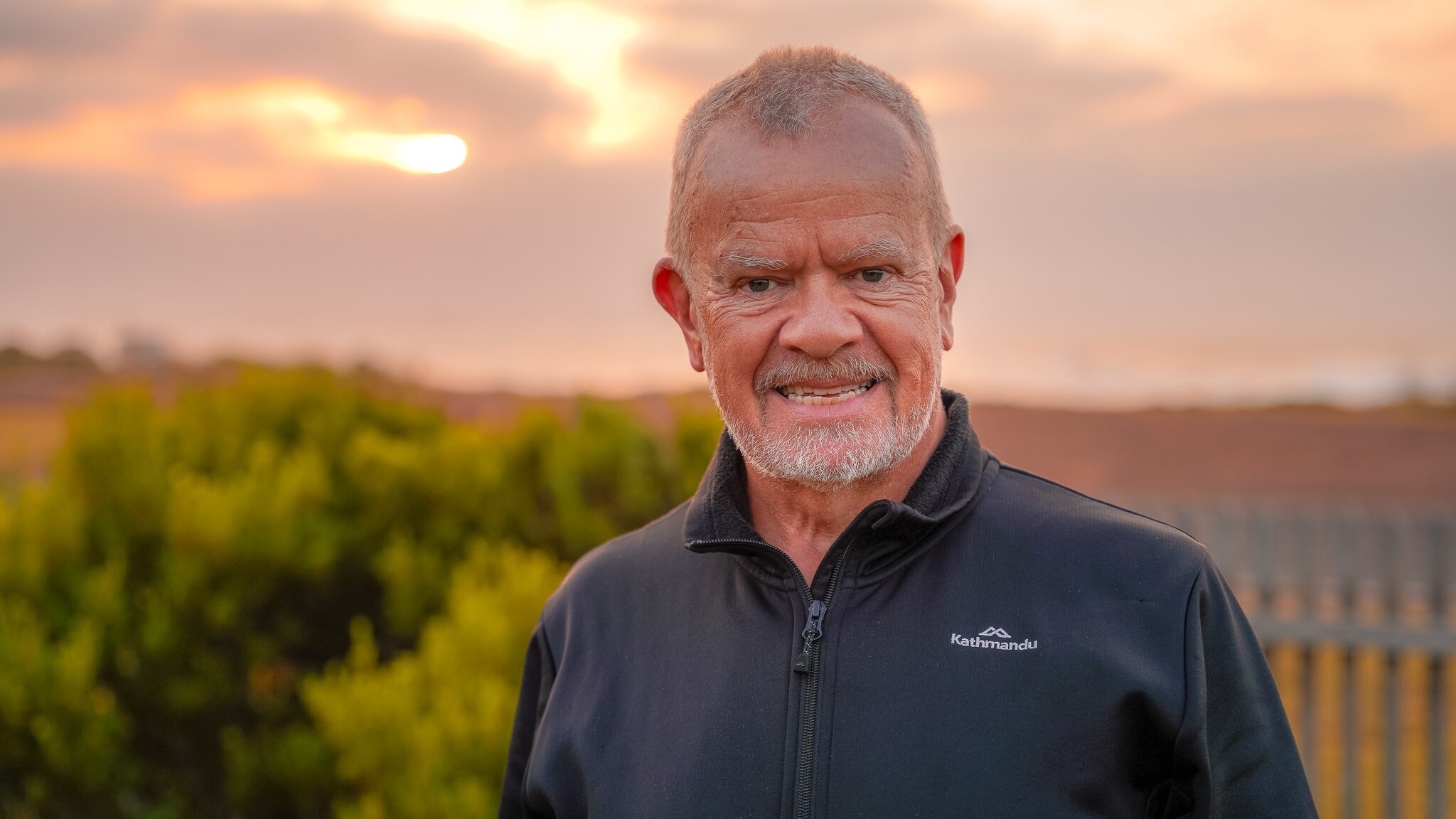A man wearing a black hiking jacket stands in a garden smiling, a pink and yellow sunrise behind him.