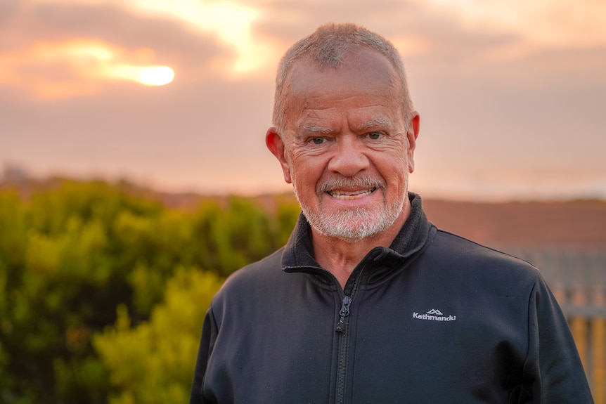 A man wearing a black hiking jacket stands in a garden smiling, a pink and yellow sunrise behind him.