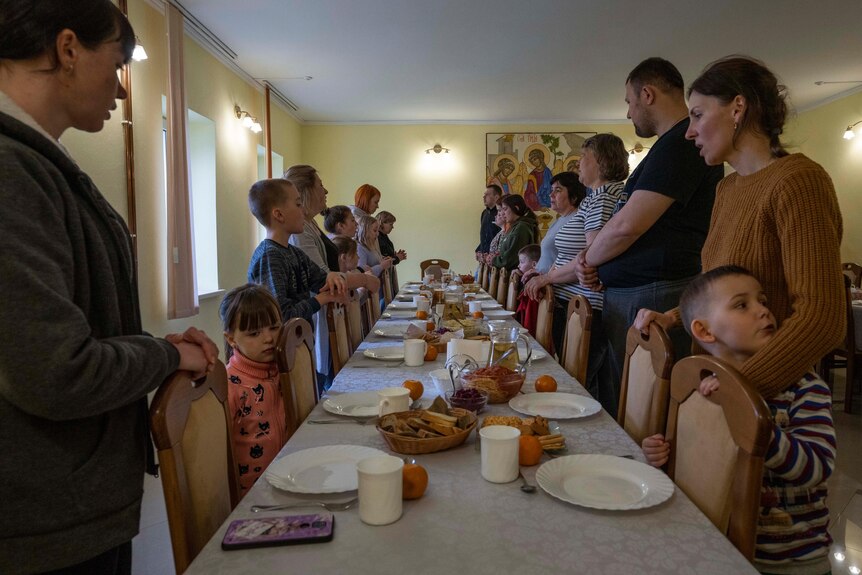 Adults and children stand either side of a long table, adorned with plates, food and cutlery.