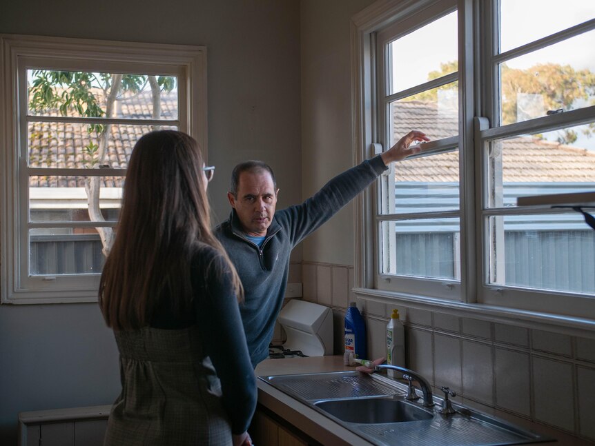 A man and woman stand in a kitchen in front of a sink, he extends a hand towards a window
