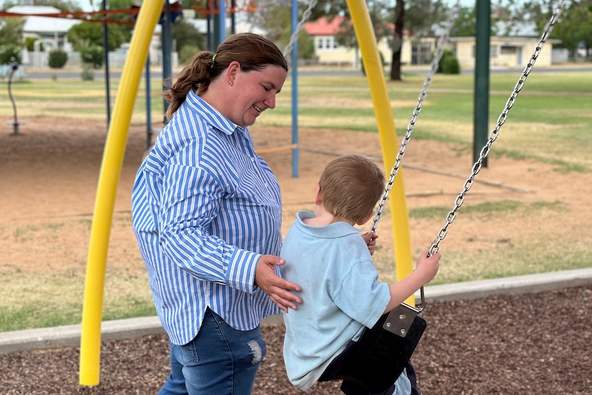 A woman stands with her hand on the back of a boy who is sitting on a swing in a playground