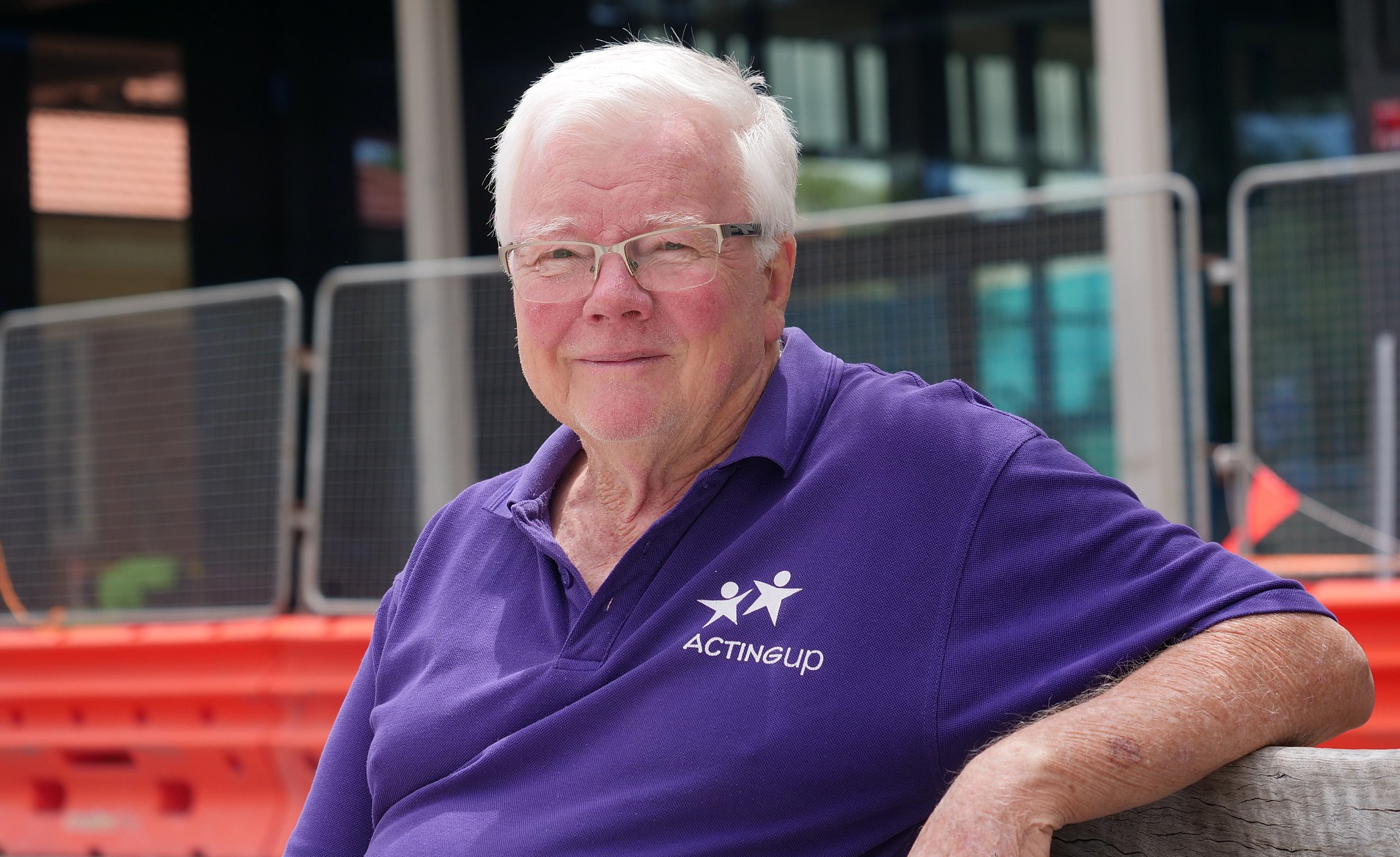 Un hombre con una camiseta de “Acting Up” sentado frente a una obra en construcción 