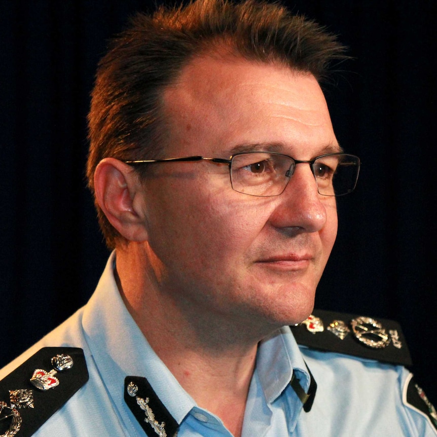 Side profile of AFP Commissioner Reece Kershaw, in a room with flags hanging behind him.