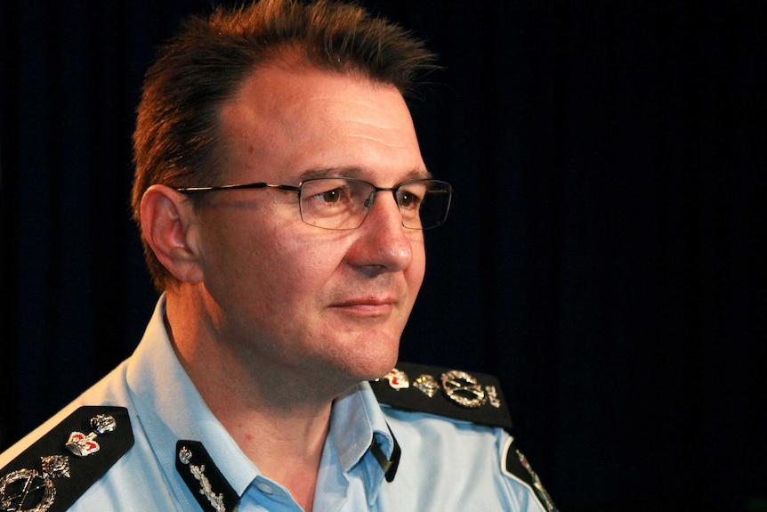 Side profile of AFP Commissioner Reece Kershaw, in a room with flags hanging behind him.