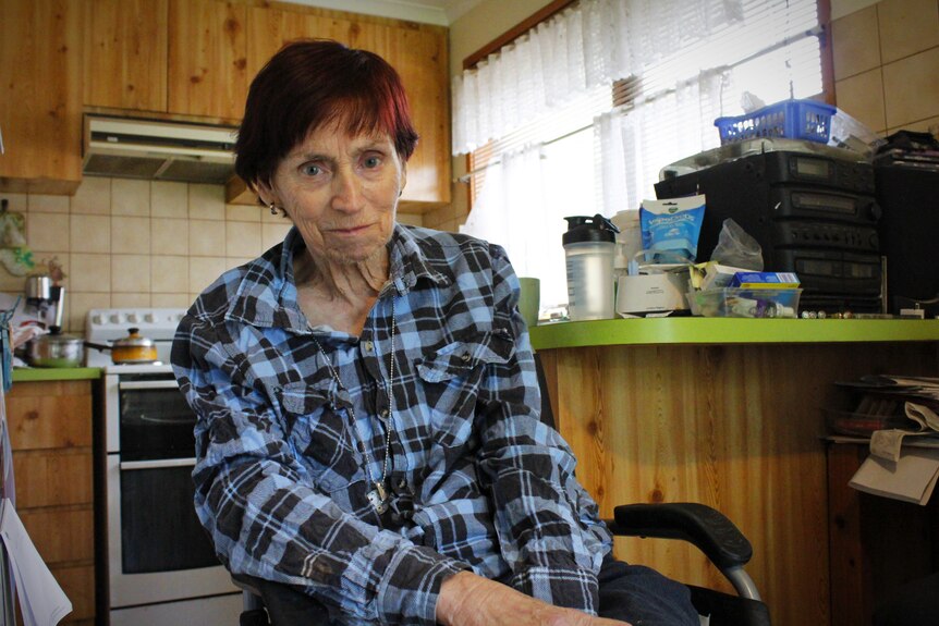 A woman in a checked blue shirt sitting in a wheelchair in her kitchen