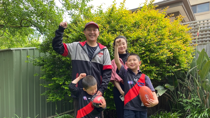You look at a family in their garden raising their firsts while wearing Essendon Football Club merch.