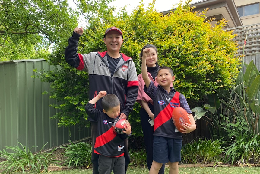You look at a family in their garden raising their firsts while wearing Essendon Football Club merch.