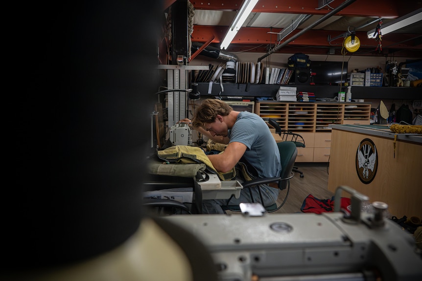 A man bends over a sewing machine in a large workroom with a bench and sewing materials stored by the wall.