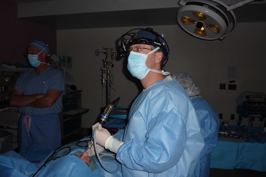 A doctor in blue scrubs standing in an operating theatre, during a procedure on a patient's nose.