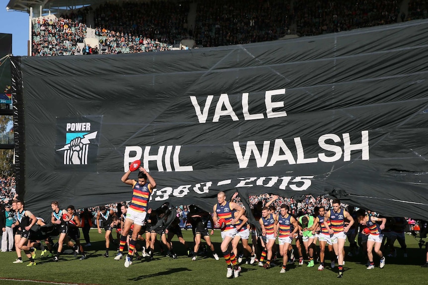 Crows and Port Adelaide players run through the banner commemorating the late Phil Walsh at Adelaide Oval