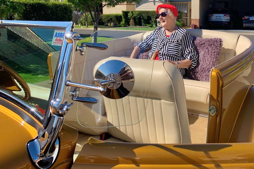 An elderly woman in a colourful top and a bright red beret and sunglasses laughs as she sits in backseat of a gold vintage car.