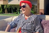 An elderly woman in a colourful top and a bright red beret and sunglasses laughs as she sits in backseat of a gold vintage car.