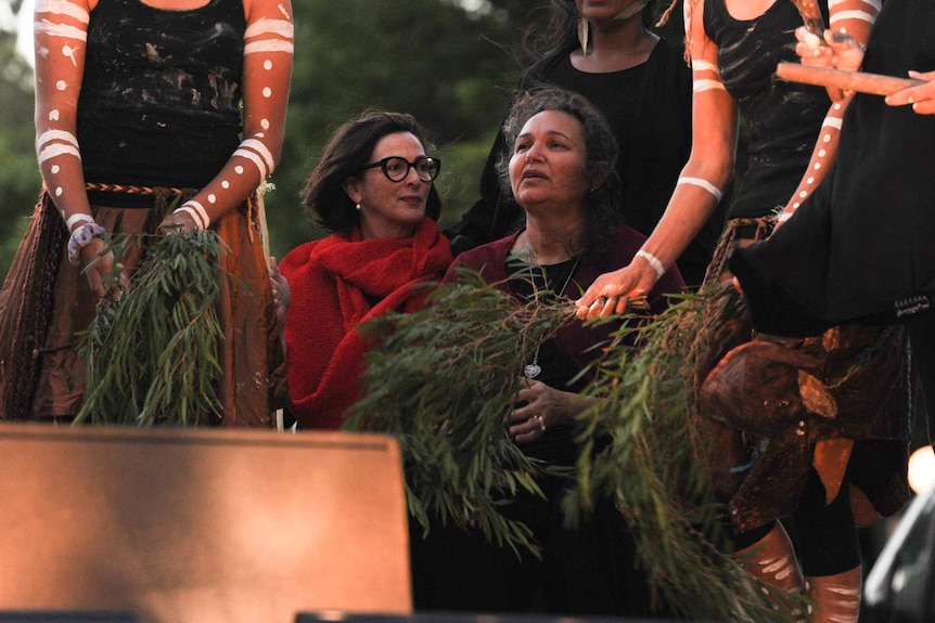 Carol Innes and Wendy Martin sit on stage with Noongar dancers holding eucalypt branches, at dusk.