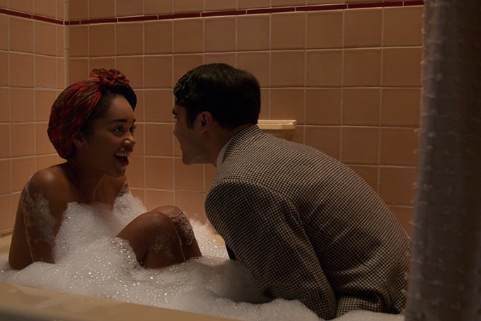 A woman with hair tied up in 1940s style headscarf sits in bubble bath looks with joy at man dressed in suit also in bath.