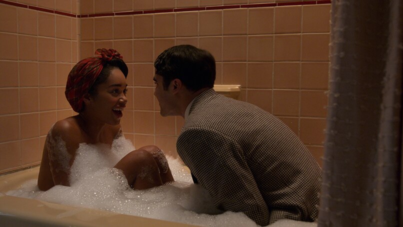 A woman with hair tied up in 1940s style headscarf sits in bubble bath looks with joy at man dressed in suit also in bath.