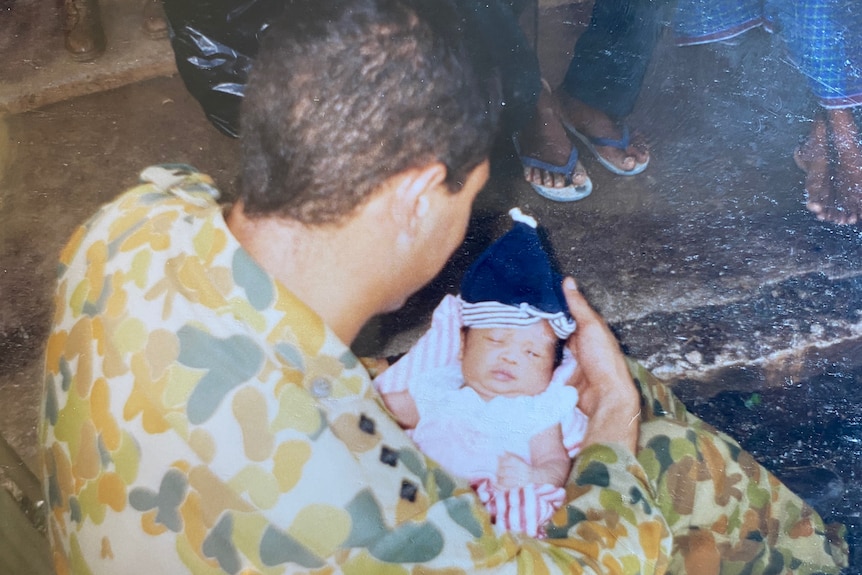 Ben in army fatigues holding a baby wrapped in blankets, taken from over ben's shoulder