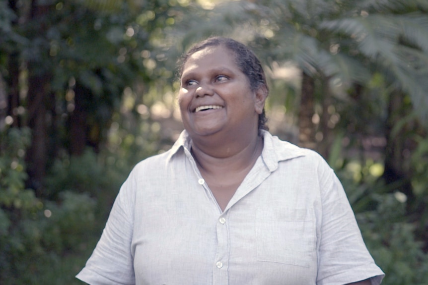 A woman in a beige shirt smiles, with a green garden behind her.