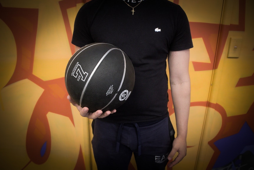 A young man in a black t-shirt holds a black basketball in one hand, standing in front of a wall of graffiti.