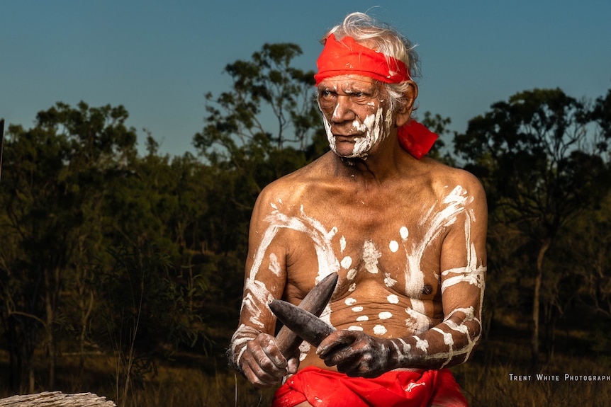 Uncle Fred pained with white dots and lines, red headband, looking to the side, holding sticks, trees behind.